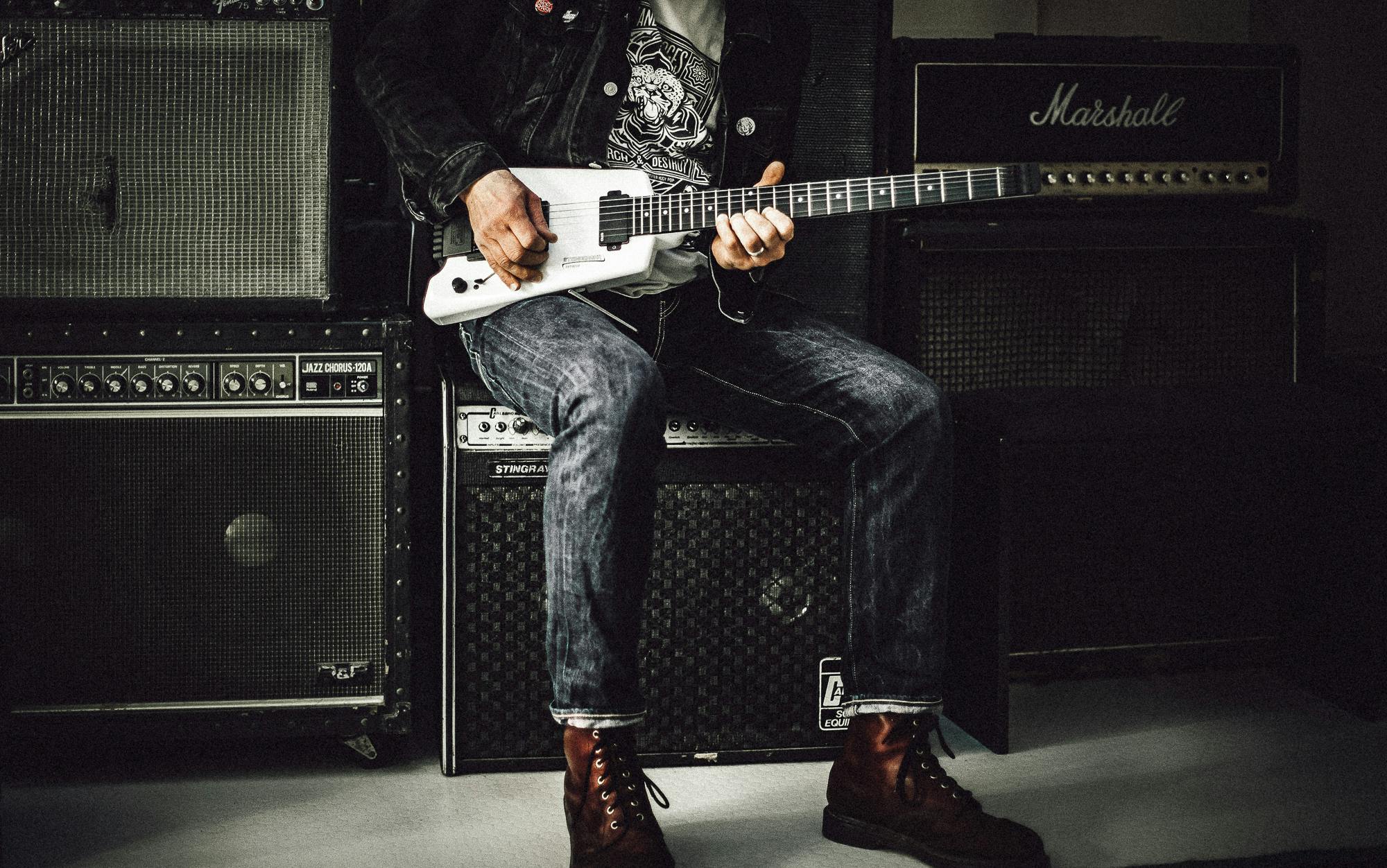 A musician plays an electric guitar in a studio, surrounded by amplifiers, showcasing rock and punk vibes.